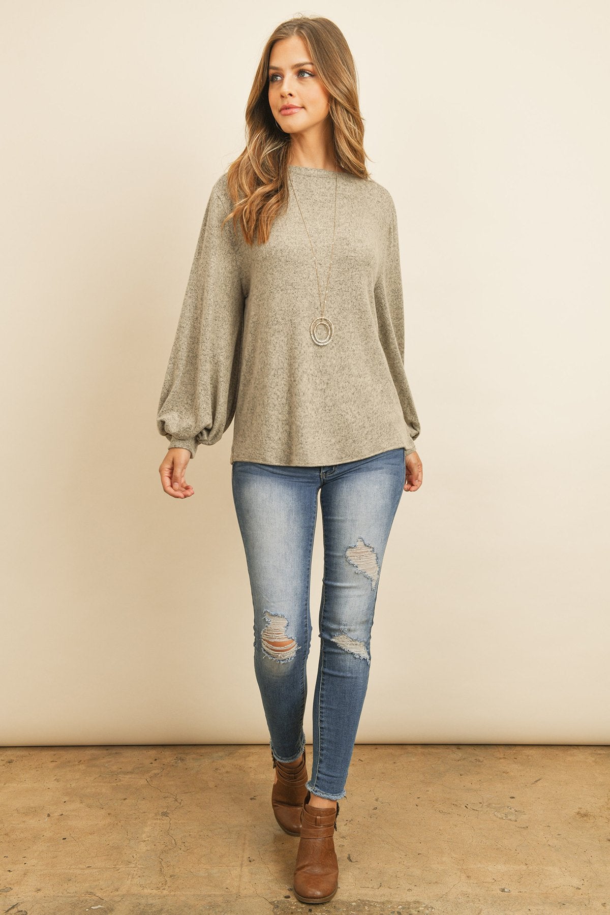 A woman models a Riah Fashion Puff Sleeved Boat Neck Two Toned Brushed Hacci Top with ripped jeans and brown boots on a concrete floor against a plain backdrop, accessorized with a long necklace.
