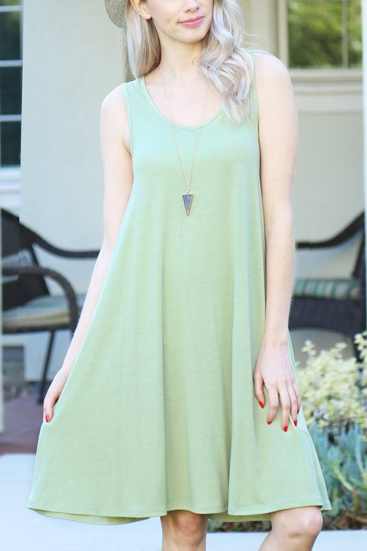 A person with long, light-colored hair wears a Sleeveless Flared Pocket Dress in light green by Riah Fashion outside, holding the dresss sides along with a triangular pendant necklace.