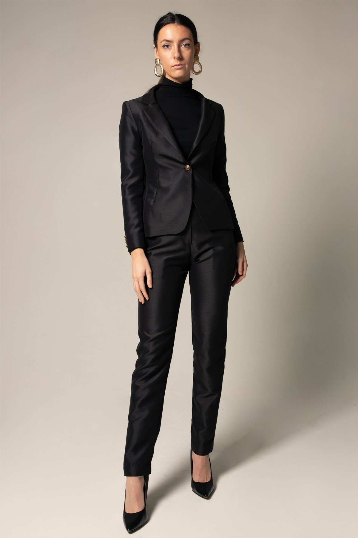 A person stands confidently in Le Réussi® Womens Blazer/Suit in Black, paired with a turtleneck and black high-heeled shoes, against a neutral background.