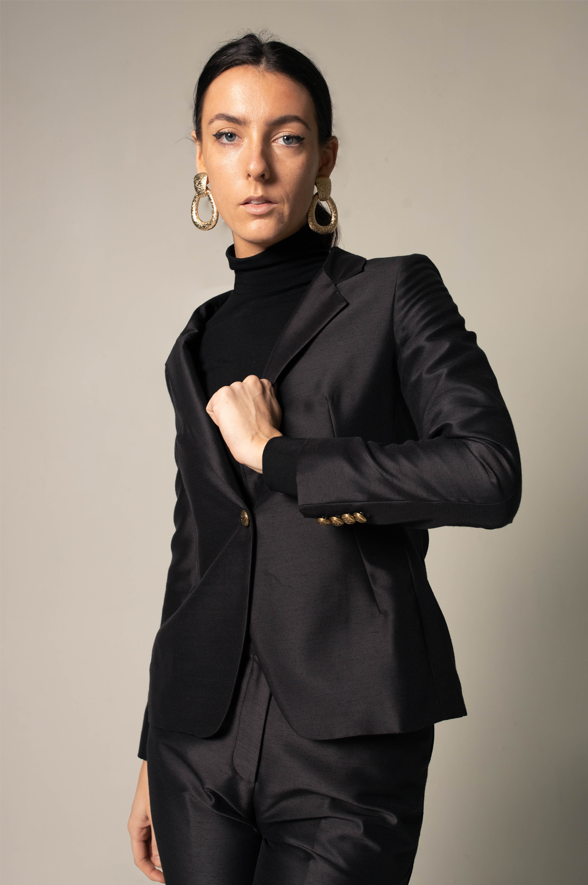 A person stands confidently in Le Réussi® Womens Blazer/Suit in Black, paired with a turtleneck and black high-heeled shoes, against a neutral background.