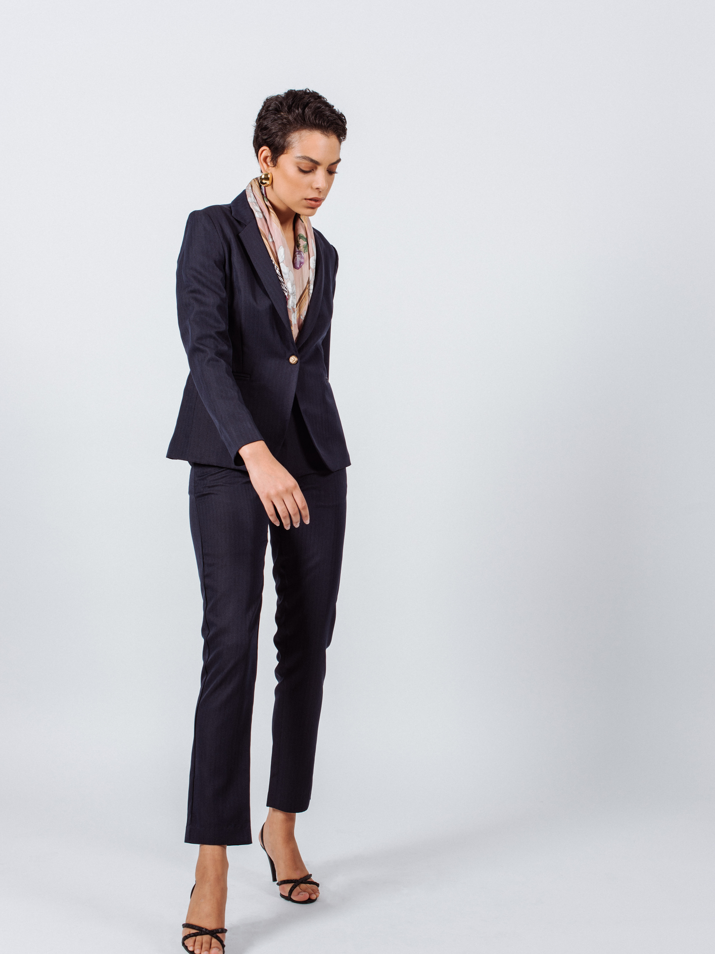 Person in Italian Wool Skinny Pants by Le Réussi®, a dark suit, heels, and a scarf stands against a plain background.