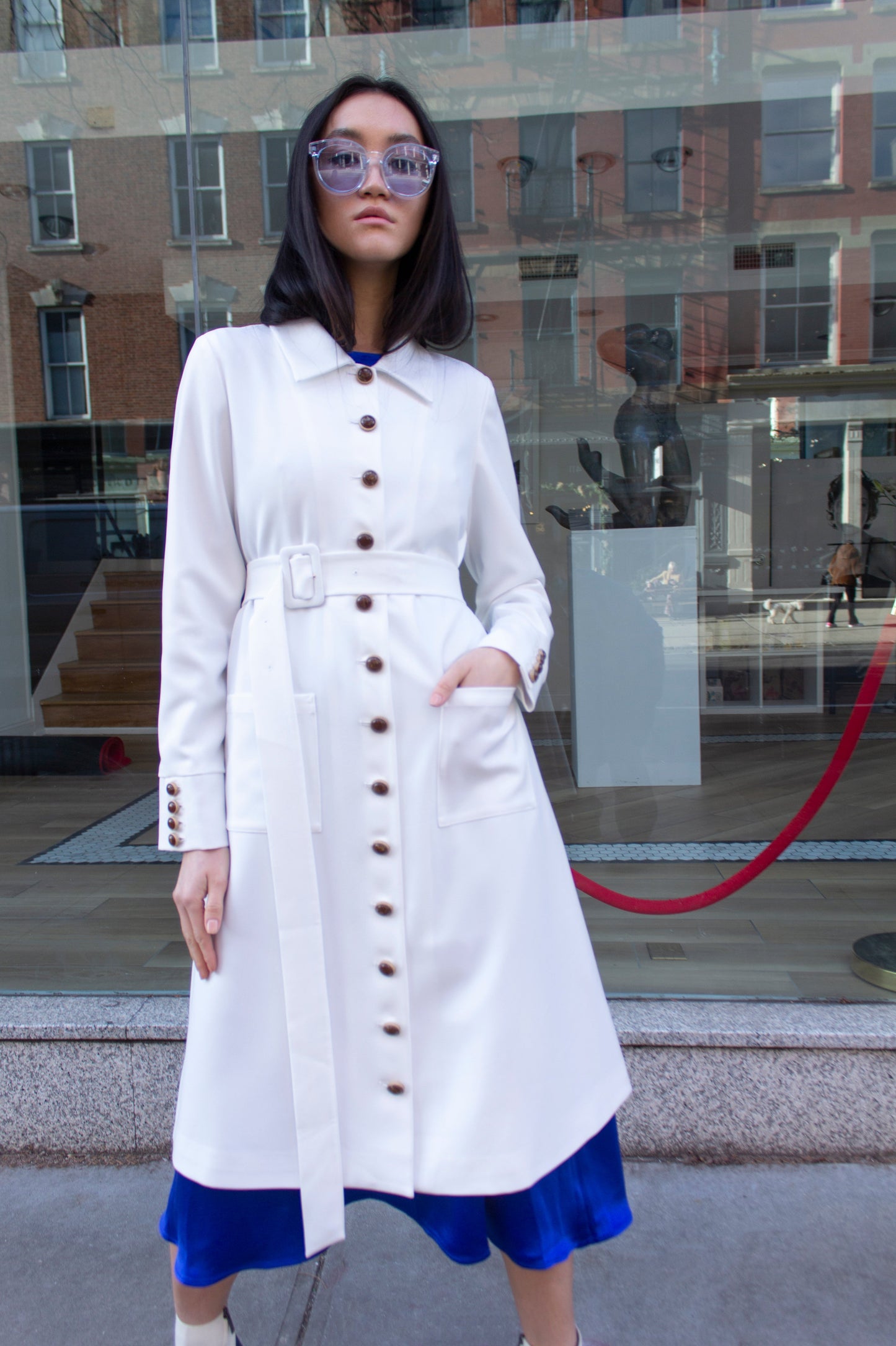 The Monique White Coat by Le Réussi® is a long-sleeve trench coat featuring a collar, button closure, matching belt, and cuff buttons.