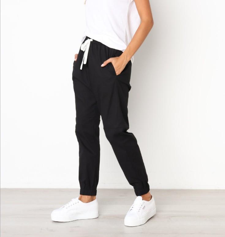 A woman models indoors in Savoy Actives Finnley Jogger Pant with a drawstring waist in black, pairing it with a crop top, her hands resting in the pockets.