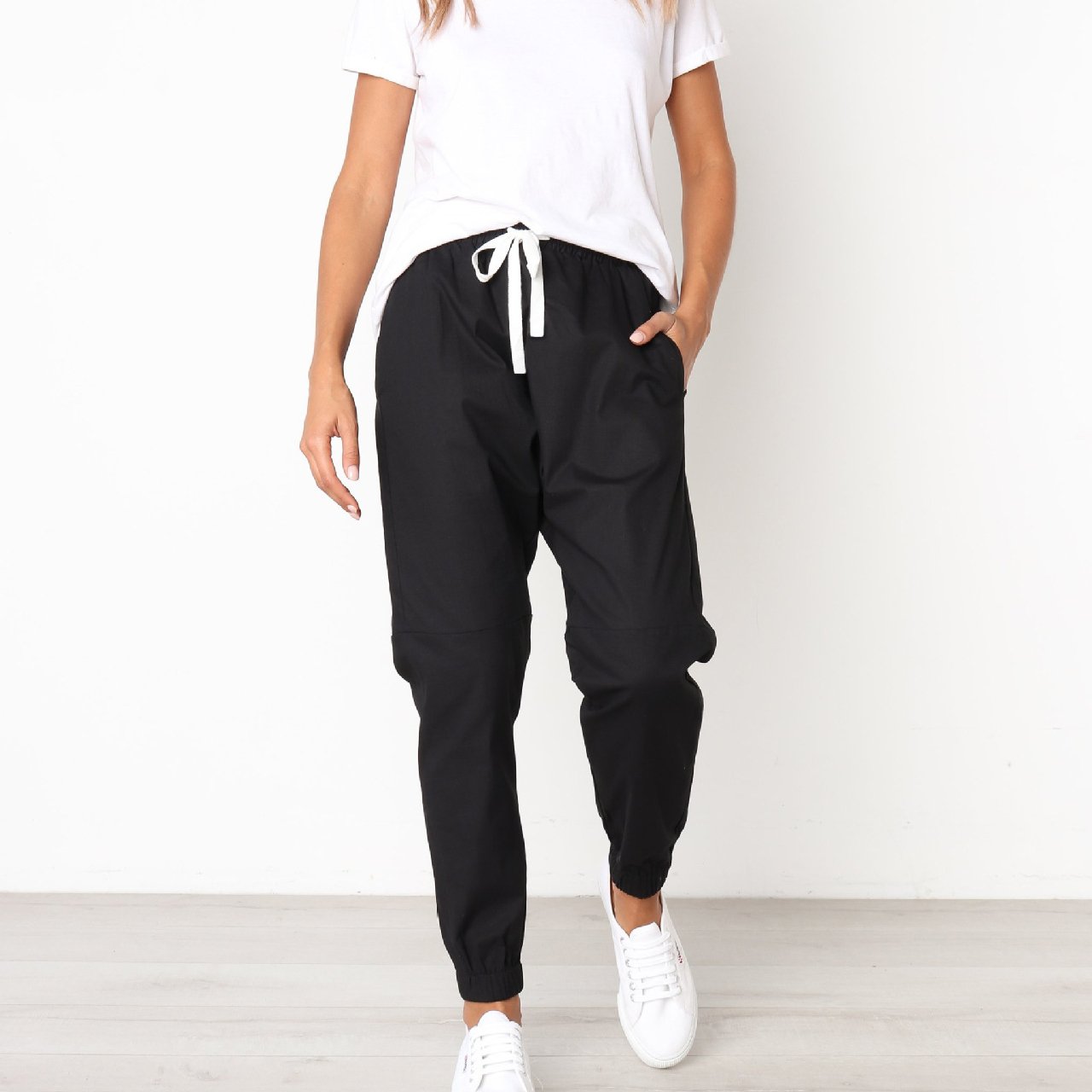 A woman models indoors in Savoy Actives Finnley Jogger Pant with a drawstring waist in black, pairing it with a crop top, her hands resting in the pockets.