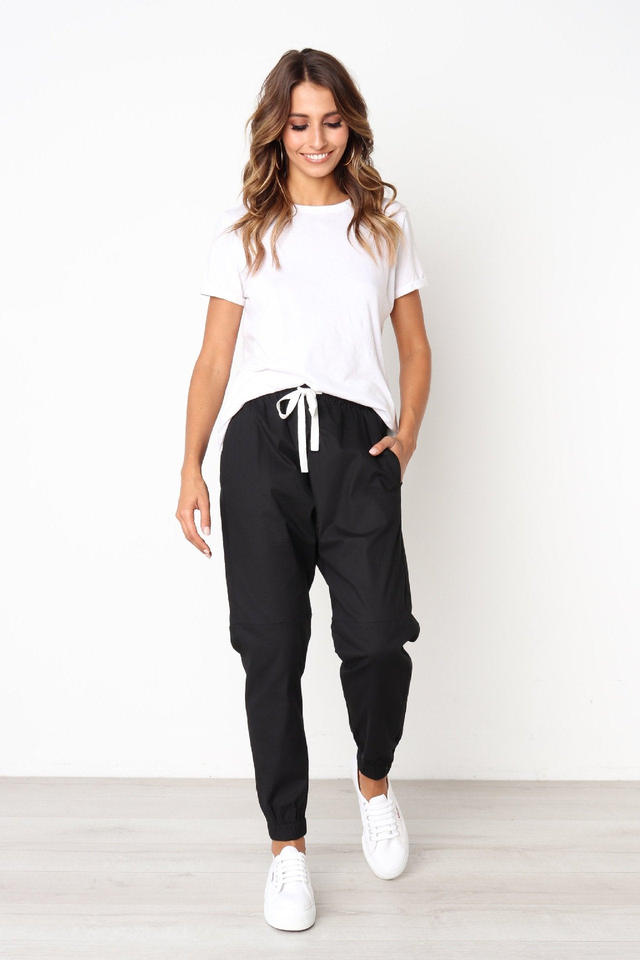 A woman models indoors in Savoy Actives Finnley Jogger Pant with a drawstring waist in black, pairing it with a crop top, her hands resting in the pockets.