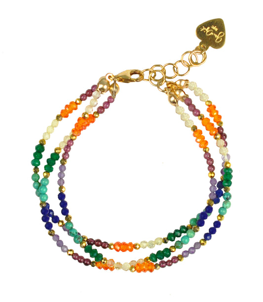 The Chakra Triple Strand Bracelet by Gena Myint is a multicolored beaded bracelet with three strands, featuring a gold clasp and chain extender.