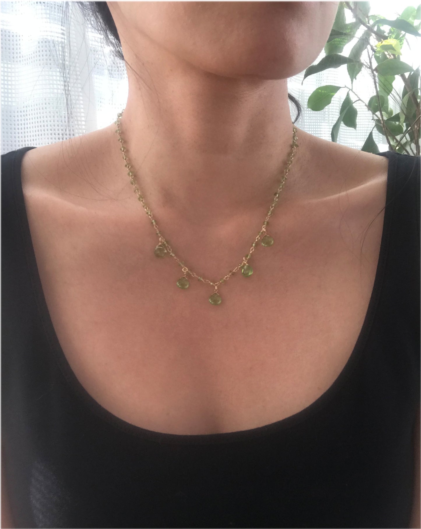 The Gena Myint Peridot Droplet Vermeil Necklace features a gold chain with small green gemstones and five larger teardrop-shaped stones.