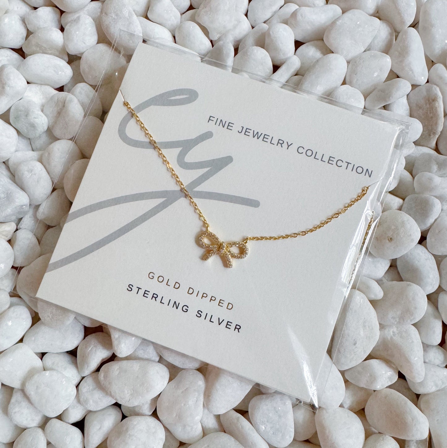 The Shine Classy Bow Sterling Silver Necklace by Ellison + Young, dipped in gold, is showcased on a card reading Fine Jewelry Collection and placed on white pebbles.