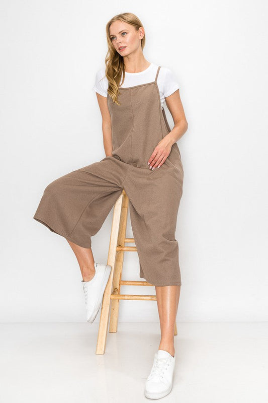 A person with long blonde hair sits on a wooden stool against a plain background, wearing Stylespects Cropped Bottom Wide Leg Oversized Jumpsuit in taupe and white sneakers.