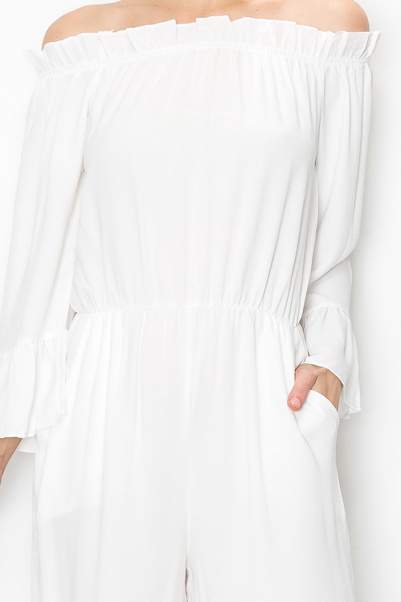 A person models Stylespects Off Shoulder Jumpsuit in white, featuring wide legs, paired with beige heels against a white backdrop.