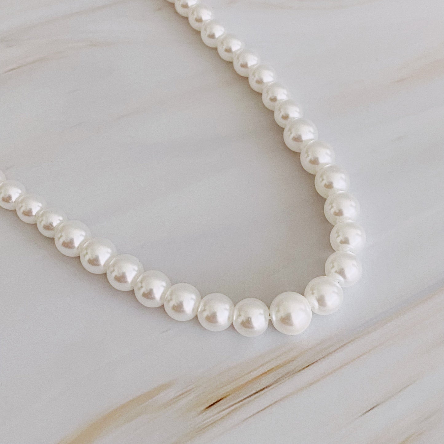 The Queen Liz Graduated Pearl Necklace by Ellison + Young features white pearls with a gold clasp, displayed on a light marble surface.