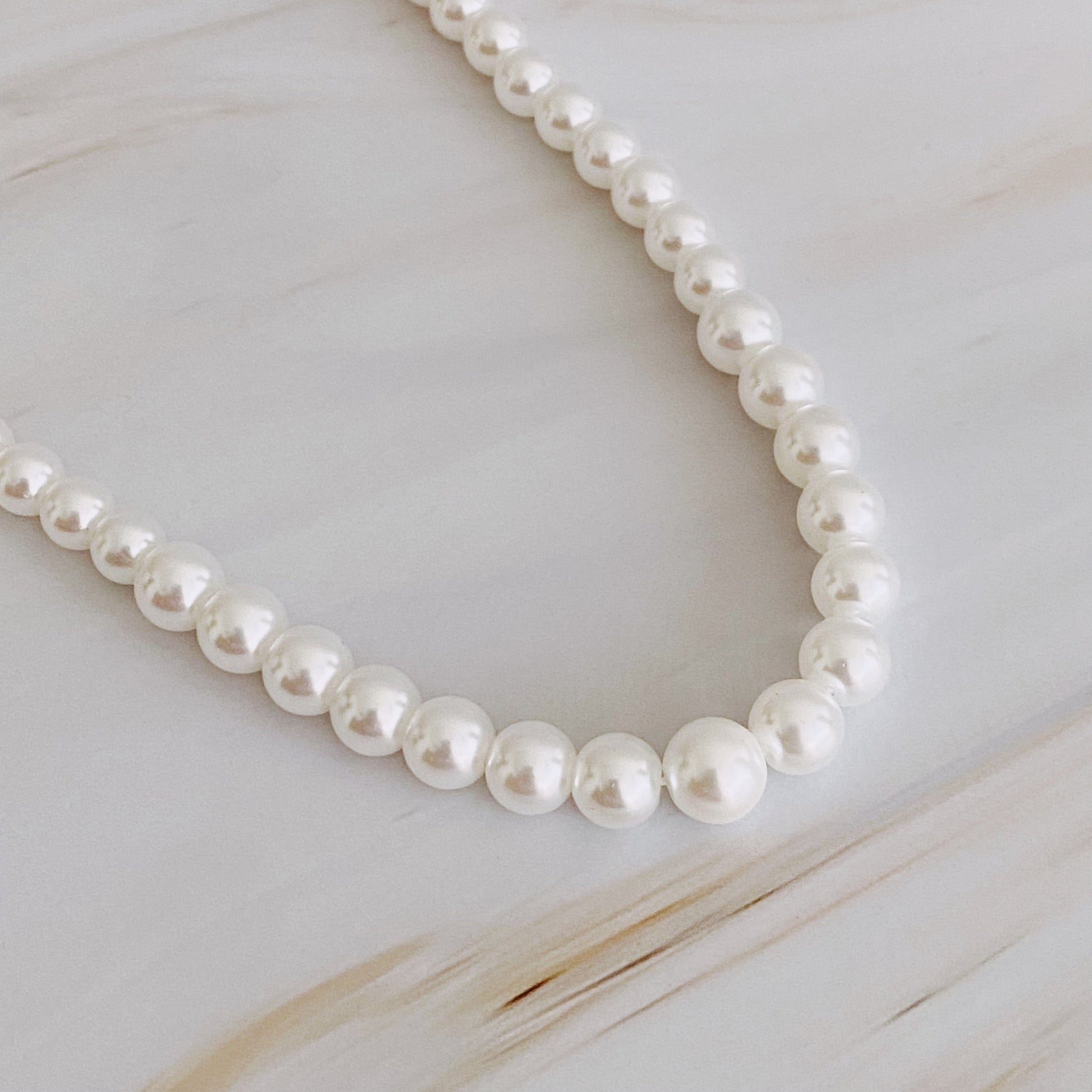 The Queen Liz Graduated Pearl Necklace by Ellison + Young features white pearls with a gold clasp, displayed on a light marble surface.