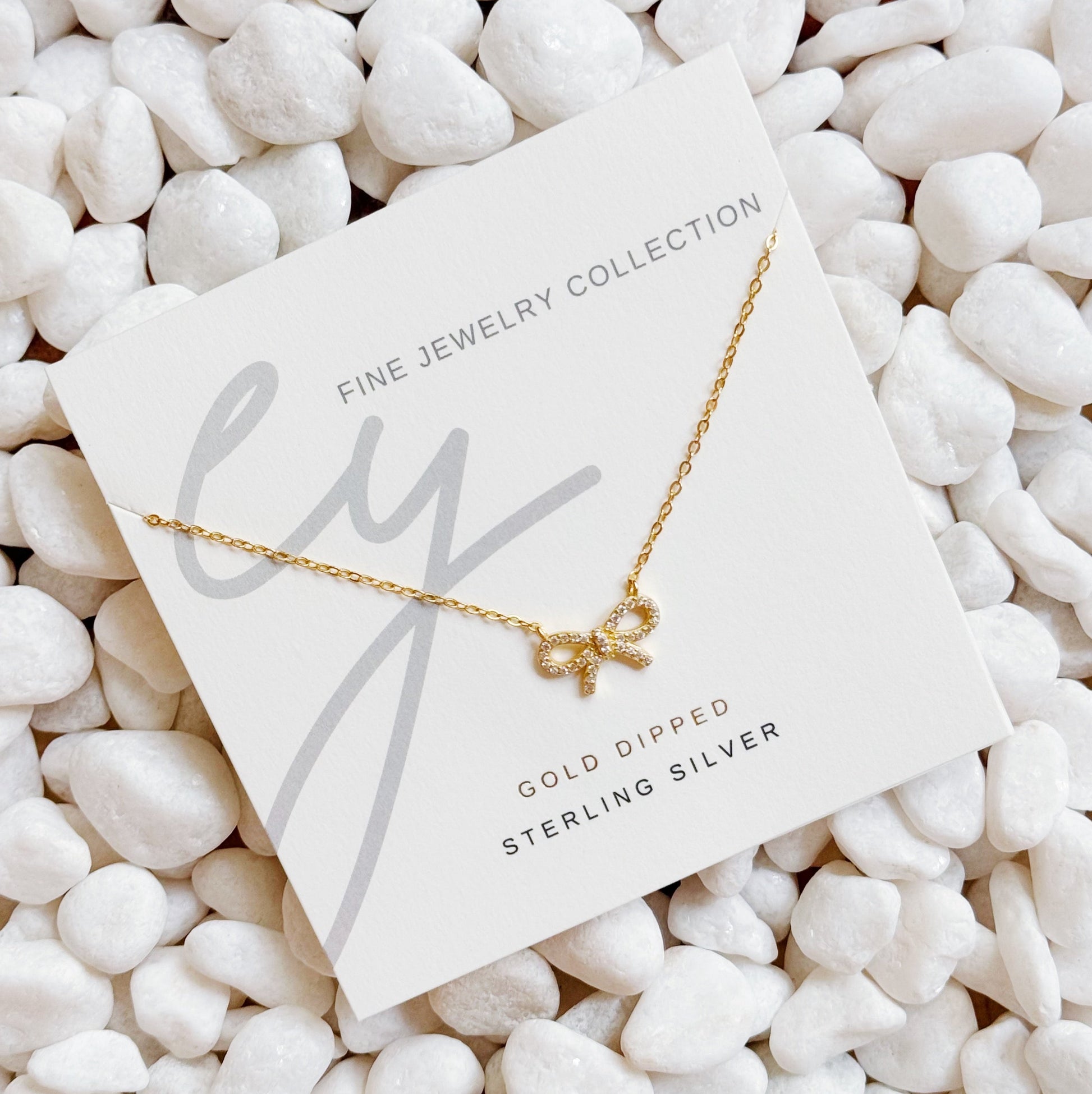 The Shine Classy Bow Sterling Silver Necklace by Ellison + Young, dipped in gold, is showcased on a card reading Fine Jewelry Collection and placed on white pebbles.