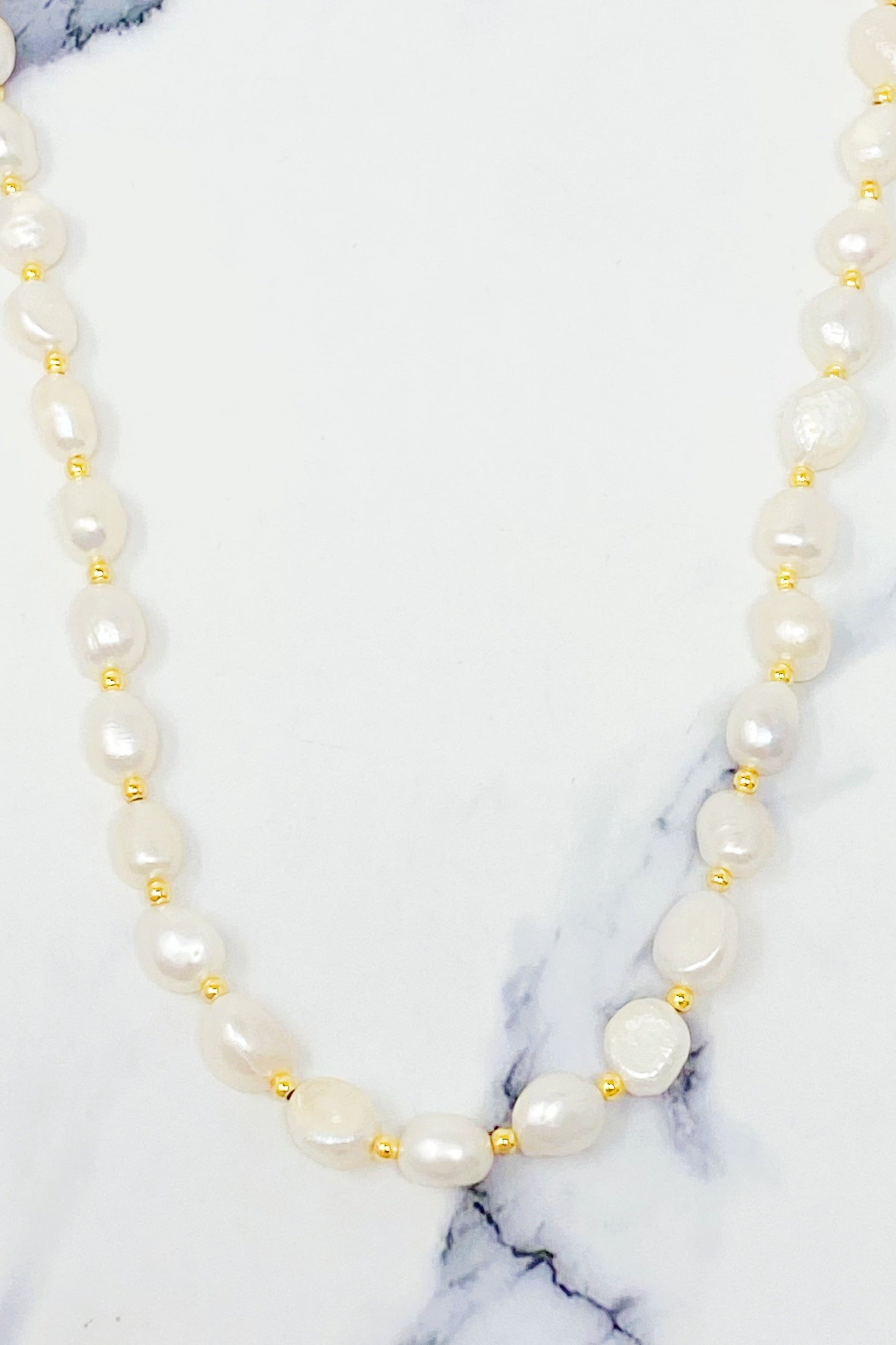 Ellison + Youngs Freshwater Pearl Necklace, featuring white pearls and small gold beads, is elegantly displayed in a curved pattern on a white marble surface.