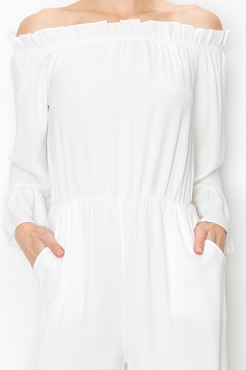 A person models Stylespects Off Shoulder Jumpsuit in white, featuring wide legs, paired with beige heels against a white backdrop.