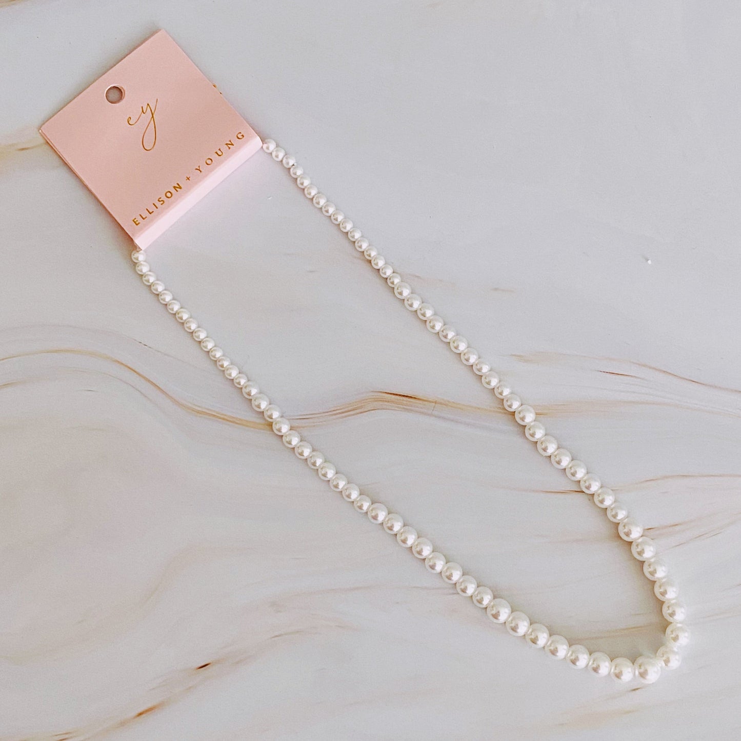 The Queen Liz Graduated Pearl Necklace by Ellison + Young features white pearls with a gold clasp, displayed on a light marble surface.