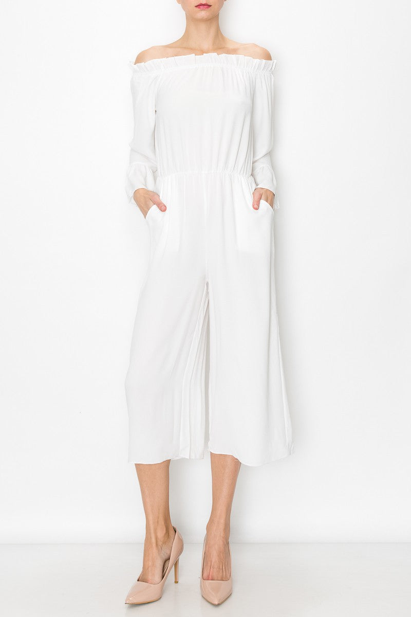 A person models Stylespects Off Shoulder Jumpsuit in white, featuring wide legs, paired with beige heels against a white backdrop.