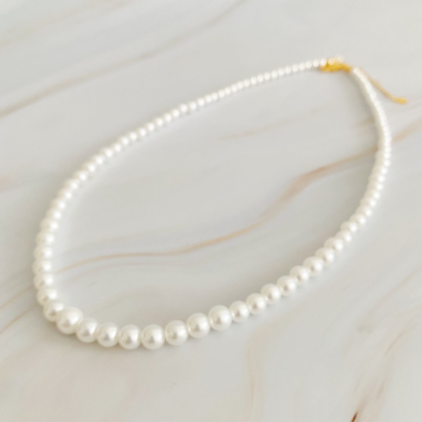 The Queen Liz Graduated Pearl Necklace by Ellison + Young features white pearls with a gold clasp, displayed on a light marble surface.