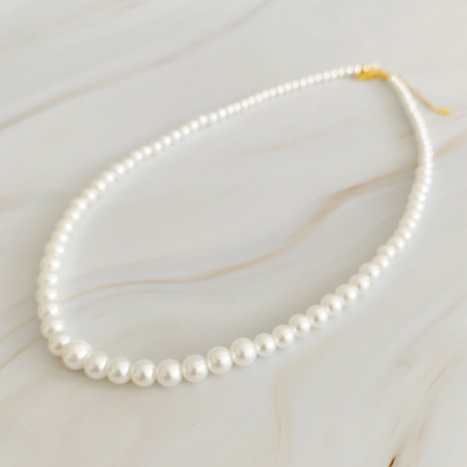 The Queen Liz Graduated Pearl Necklace by Ellison + Young features white pearls with a gold clasp, displayed on a light marble surface.