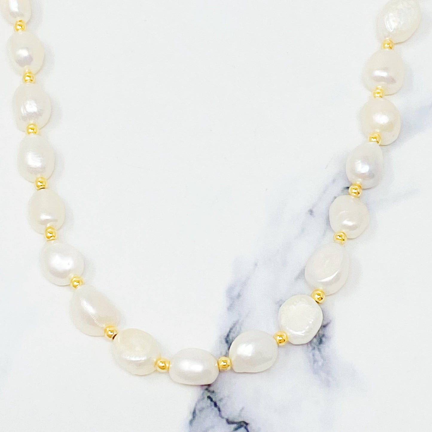 Ellison + Youngs Freshwater Pearl Necklace, featuring white pearls and small gold beads, is elegantly displayed in a curved pattern on a white marble surface.