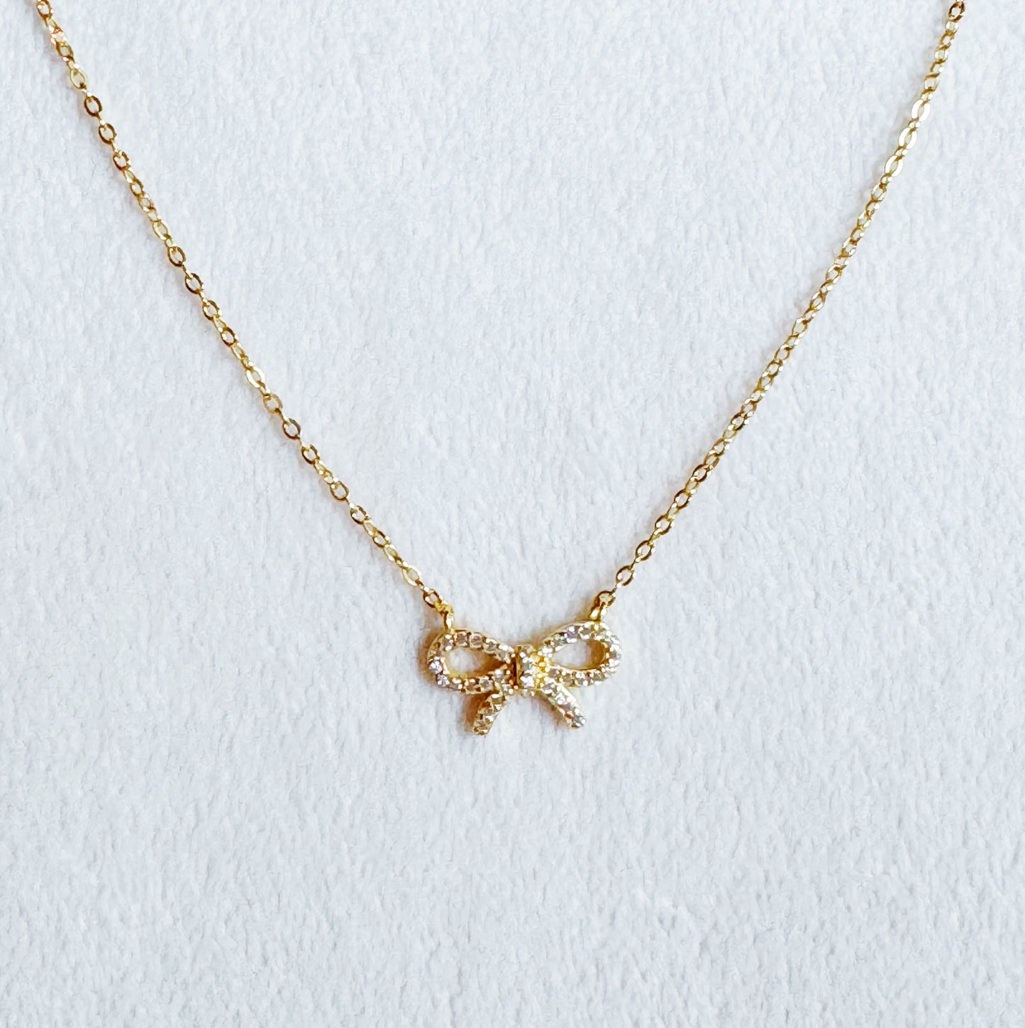 The Shine Classy Bow Sterling Silver Necklace by Ellison + Young, dipped in gold, is showcased on a card reading Fine Jewelry Collection and placed on white pebbles.