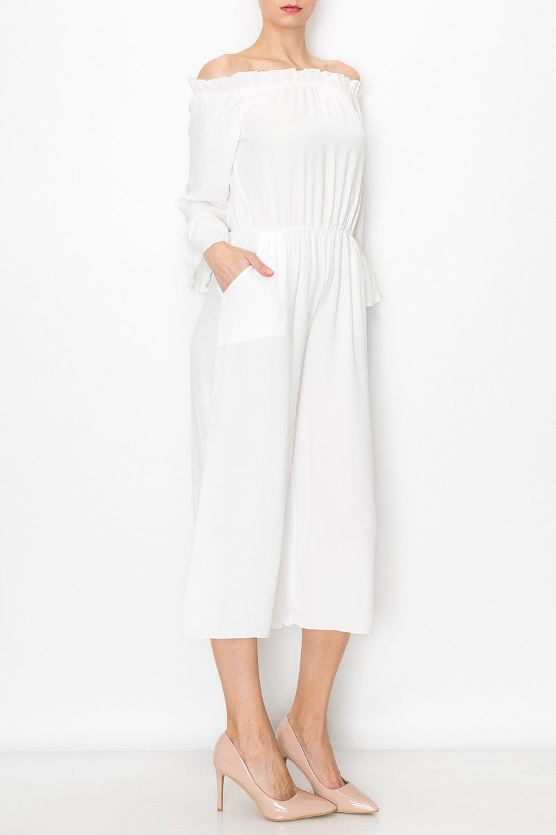 A person models Stylespects Off Shoulder Jumpsuit in white, featuring wide legs, paired with beige heels against a white backdrop.