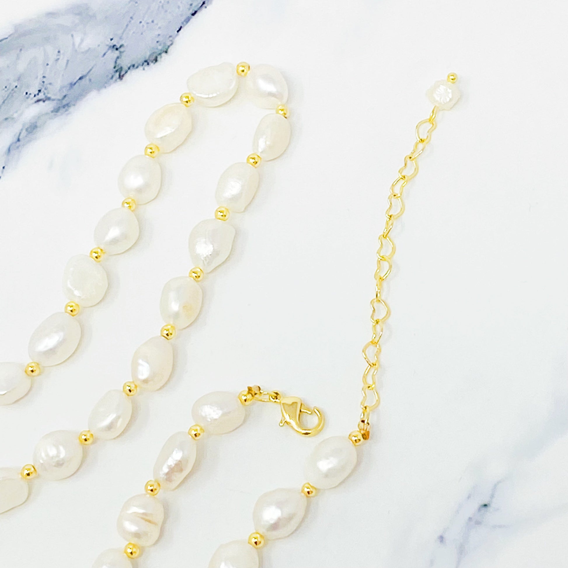 Ellison + Youngs Freshwater Pearl Necklace, featuring white pearls and small gold beads, is elegantly displayed in a curved pattern on a white marble surface.