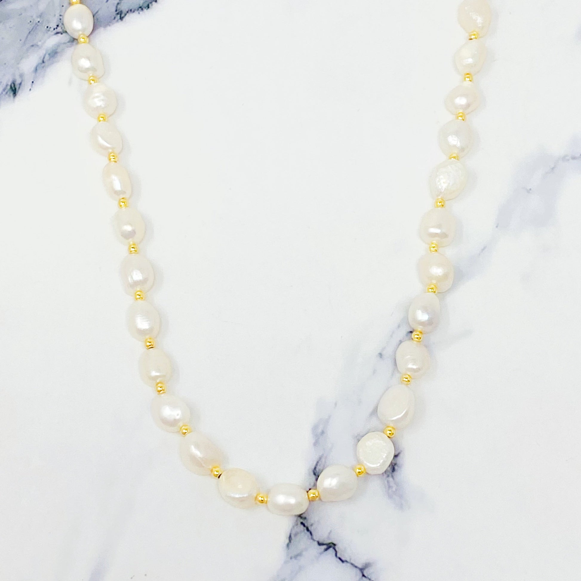 Ellison + Youngs Freshwater Pearl Necklace, featuring white pearls and small gold beads, is elegantly displayed in a curved pattern on a white marble surface.