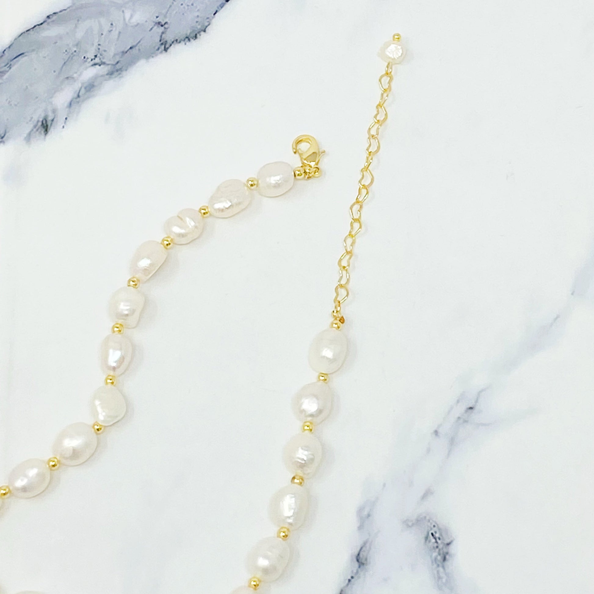 Ellison + Youngs Freshwater Pearl Necklace, featuring white pearls and small gold beads, is elegantly displayed in a curved pattern on a white marble surface.