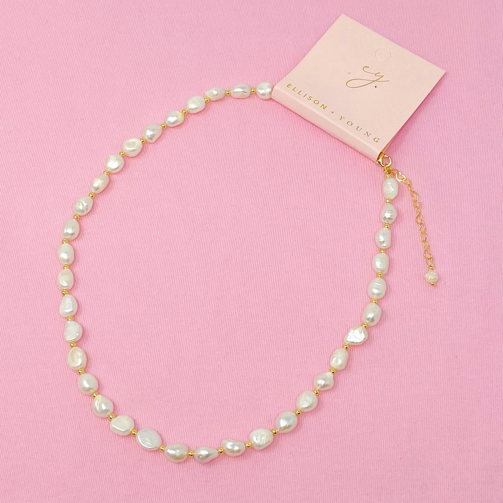 Ellison + Youngs Freshwater Pearl Necklace, featuring white pearls and small gold beads, is elegantly displayed in a curved pattern on a white marble surface.