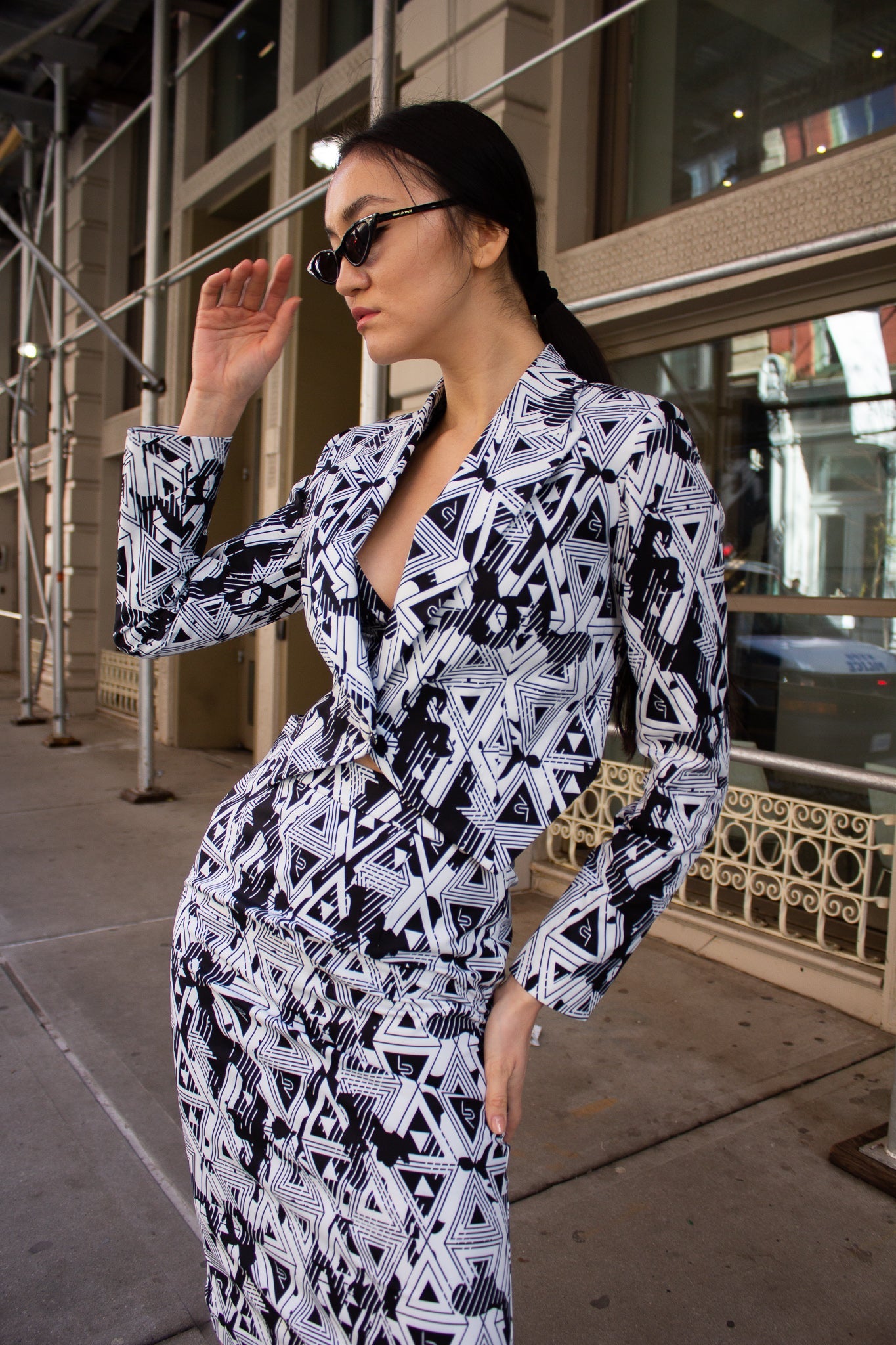 A person wearing the Amour Geometric Blazer by Le Réussi® paired with sunglasses stands on a city street.