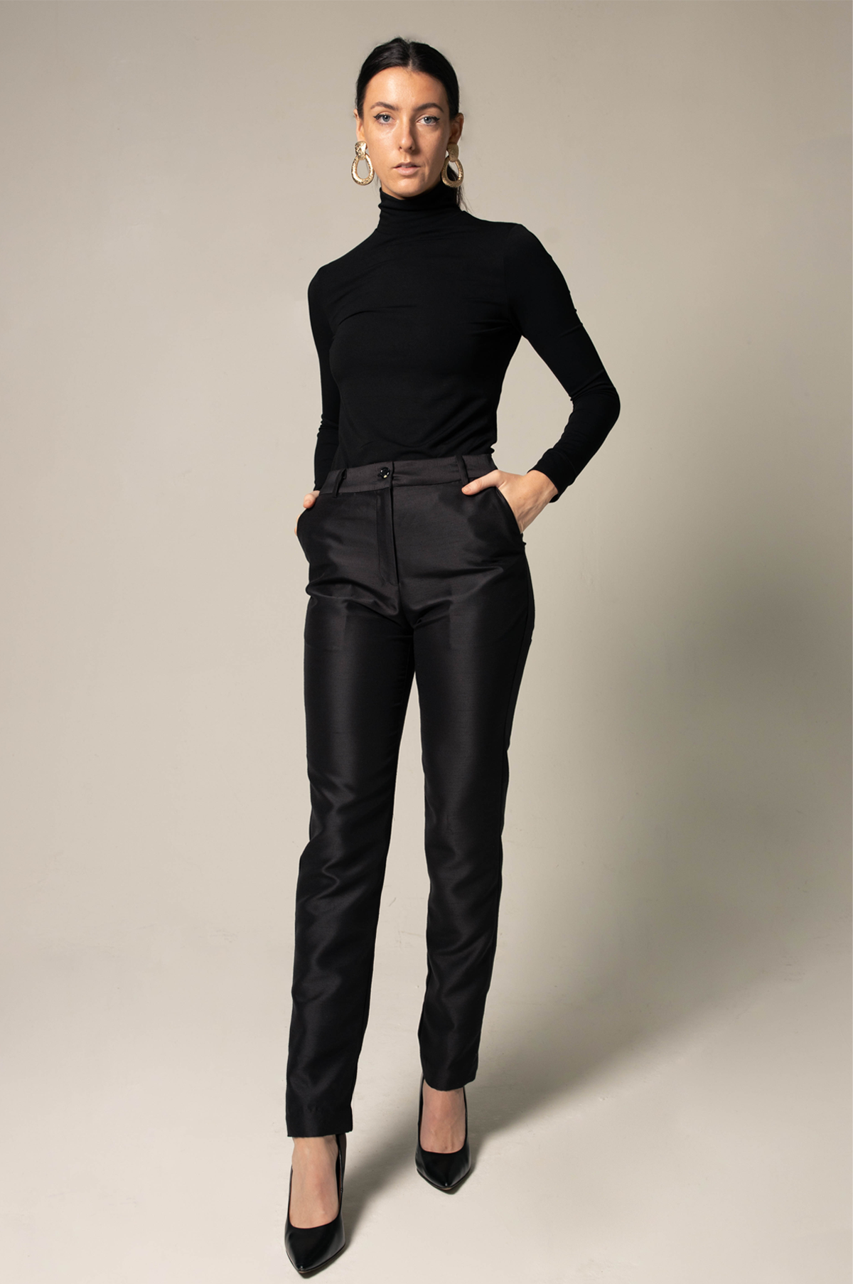 A confident individual in a black turtleneck and Le Réussi® Elegant Skinny Pants in Black stands with hands in pockets against a neutral backdrop, complemented by hoop earrings and black heels.