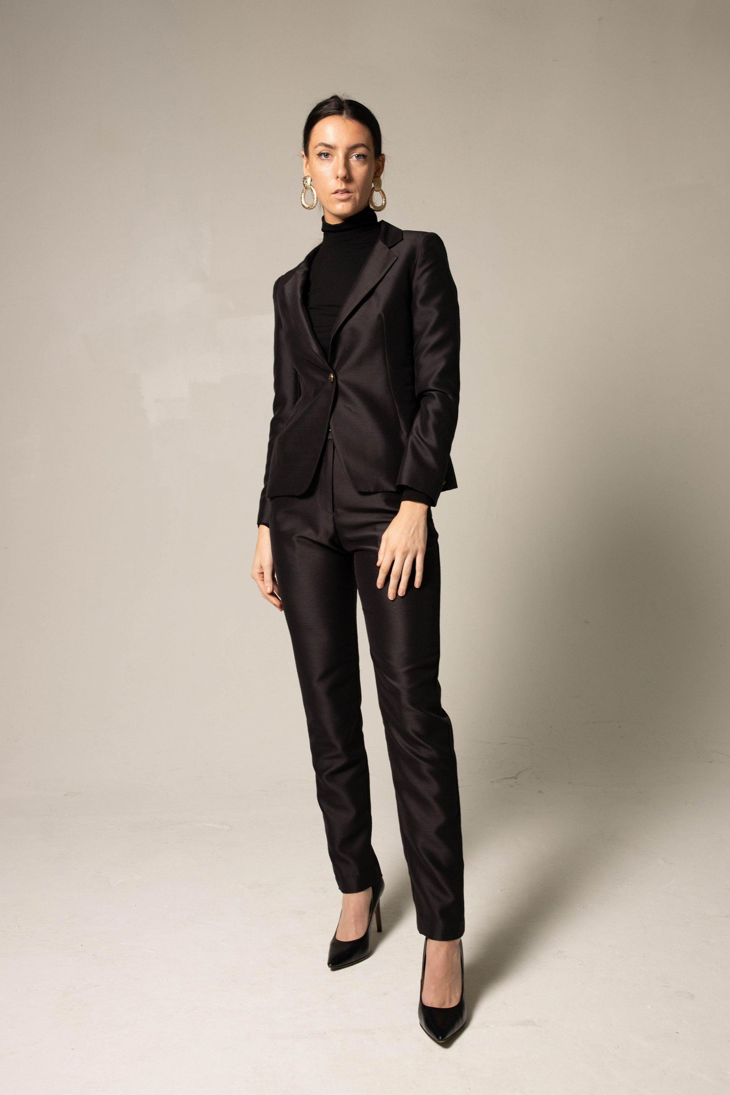 A confident individual in a black turtleneck and Le Réussi® Elegant Skinny Pants in Black stands with hands in pockets against a neutral backdrop, complemented by hoop earrings and black heels.