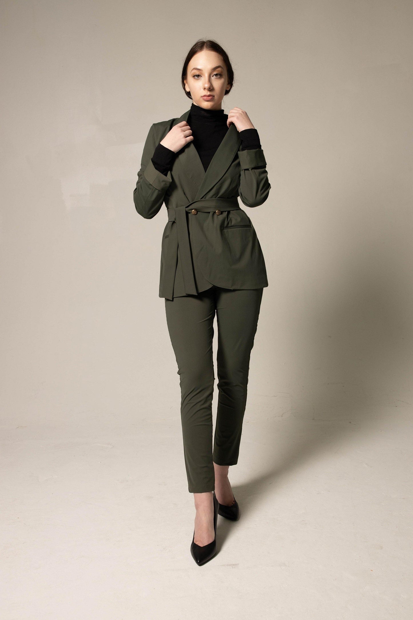 A woman wearing Le Réussi® Womens Olive Blazer With Front Buttons and a black turtleneck gazes upwards with her hands in her pockets against a neutral background.