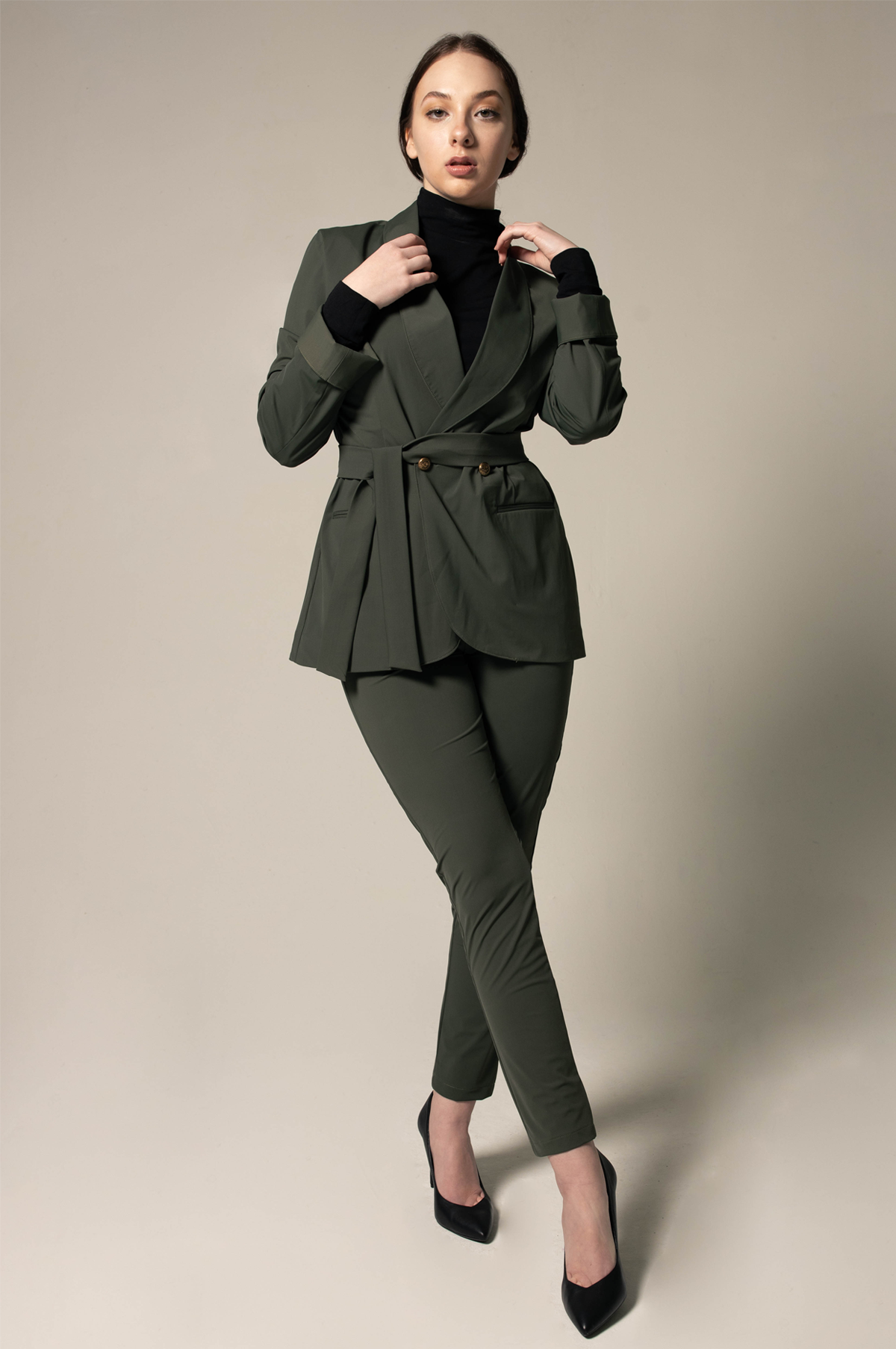 A woman wearing Le Réussi® Womens Olive Blazer With Front Buttons and a black turtleneck gazes upwards with her hands in her pockets against a neutral background.