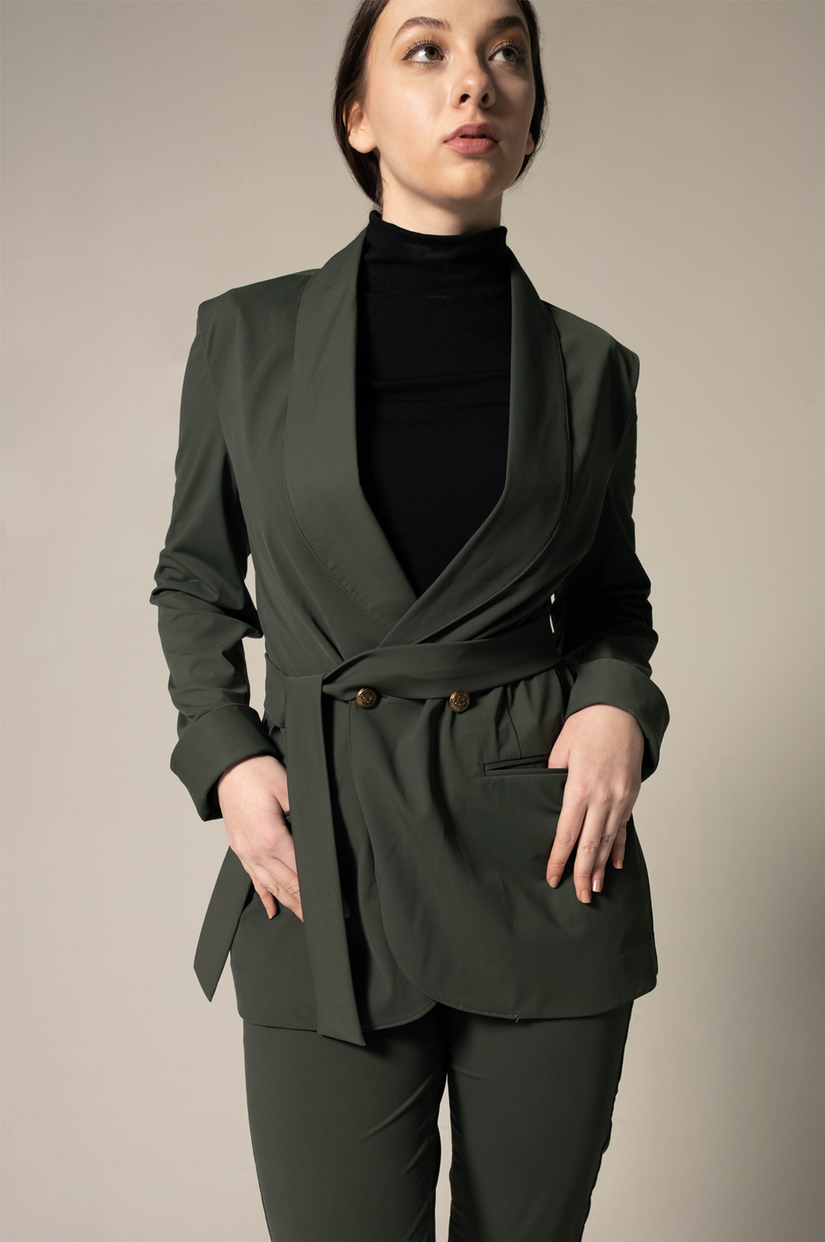 A woman wearing Le Réussi® Womens Olive Blazer With Front Buttons and a black turtleneck gazes upwards with her hands in her pockets against a neutral background.