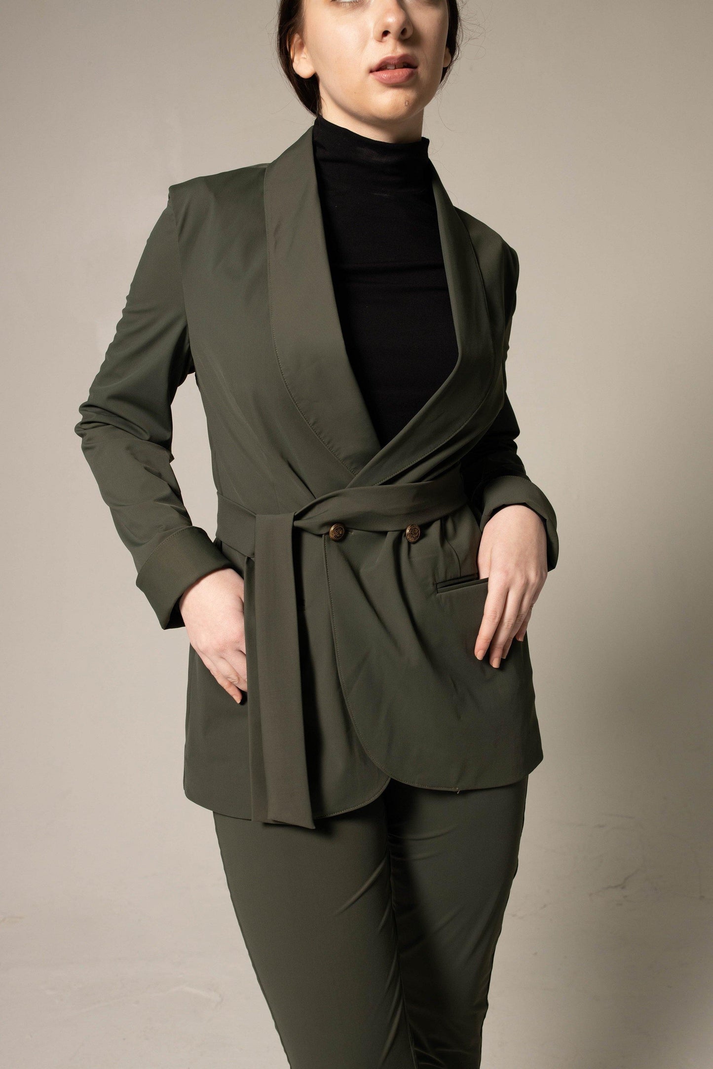 A woman wearing Le Réussi® Womens Olive Blazer With Front Buttons and a black turtleneck gazes upwards with her hands in her pockets against a neutral background.