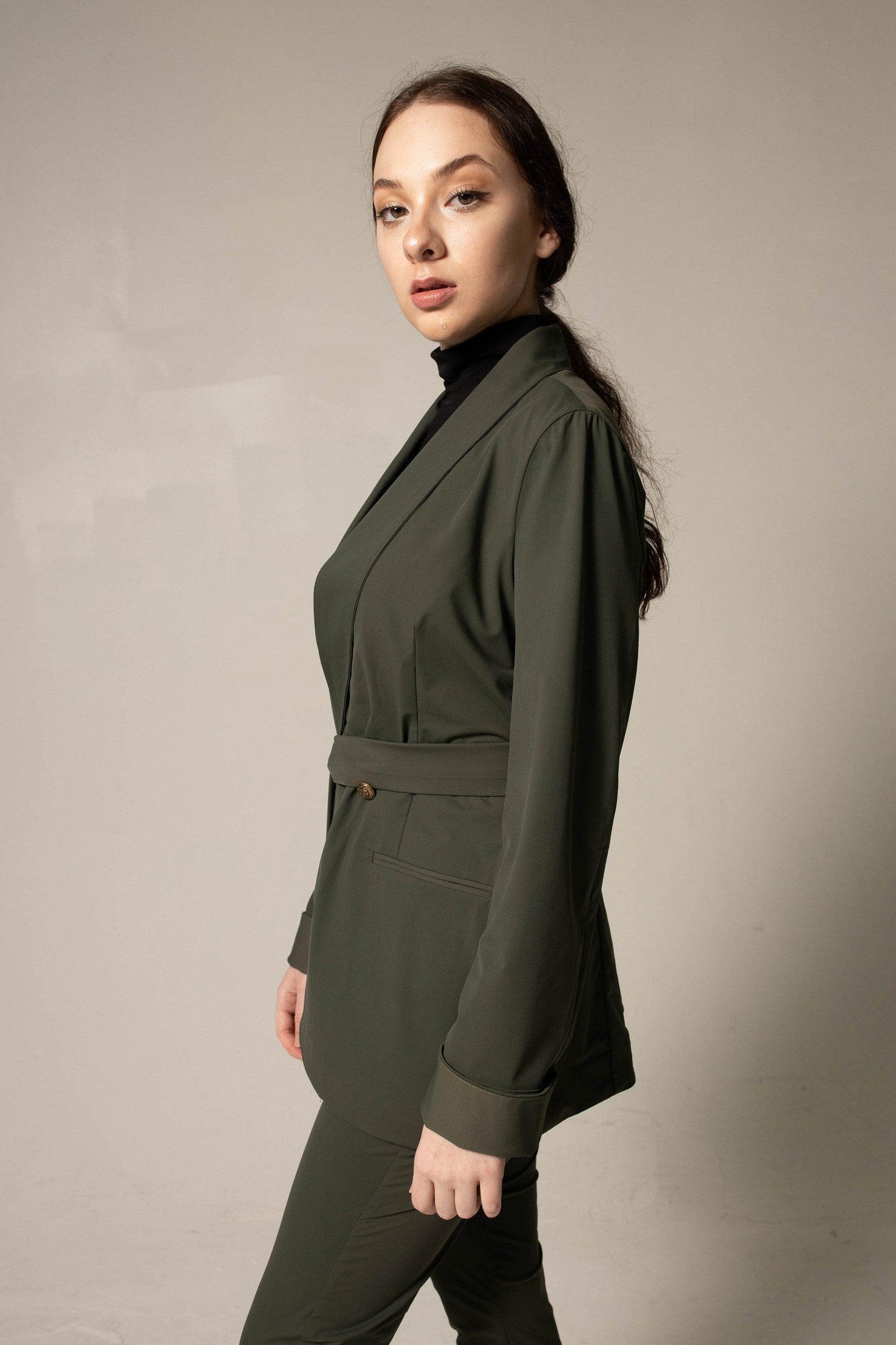 A woman wearing Le Réussi® Womens Olive Blazer With Front Buttons and a black turtleneck gazes upwards with her hands in her pockets against a neutral background.