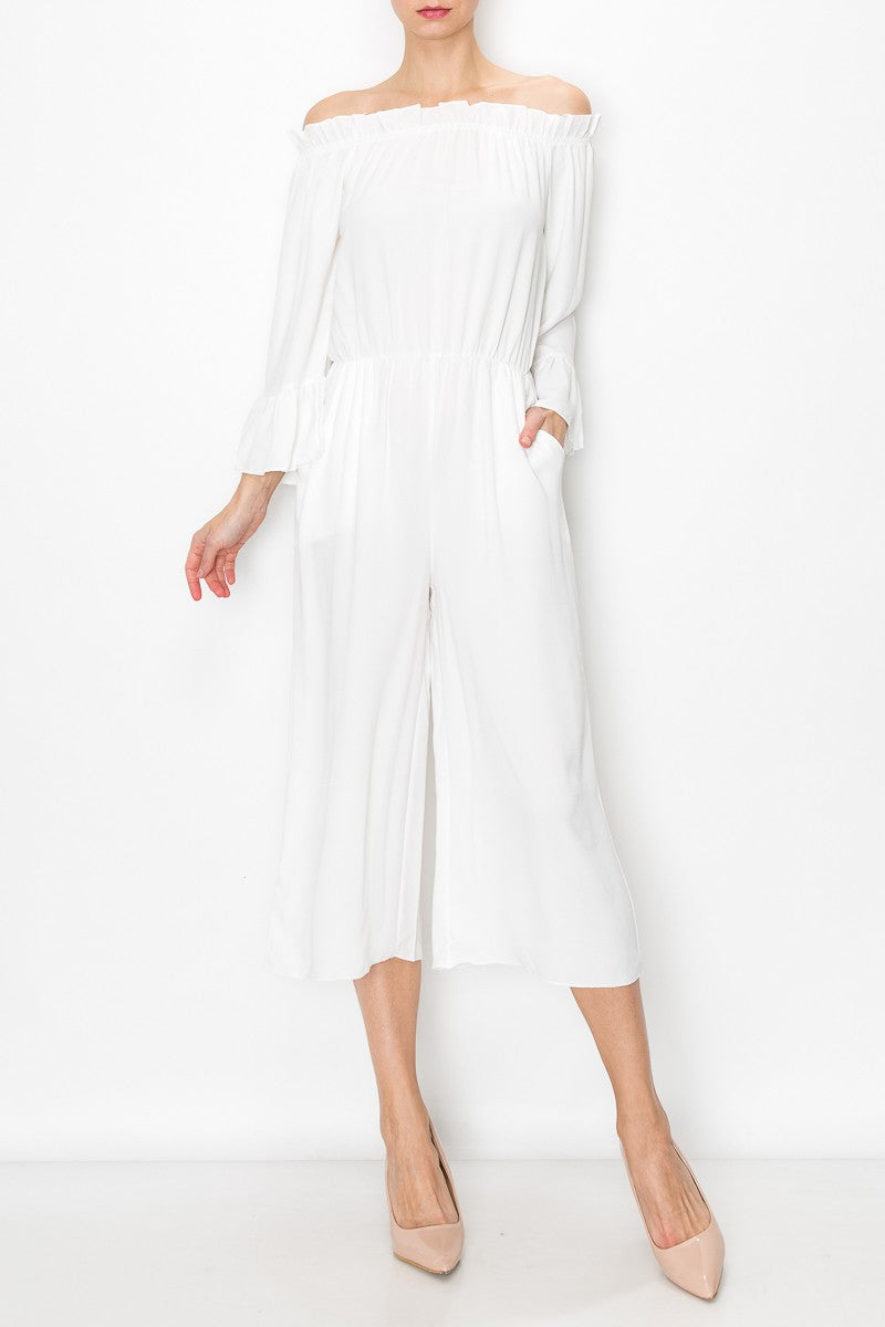 A person models Stylespects Off Shoulder Jumpsuit in white, featuring wide legs, paired with beige heels against a white backdrop.