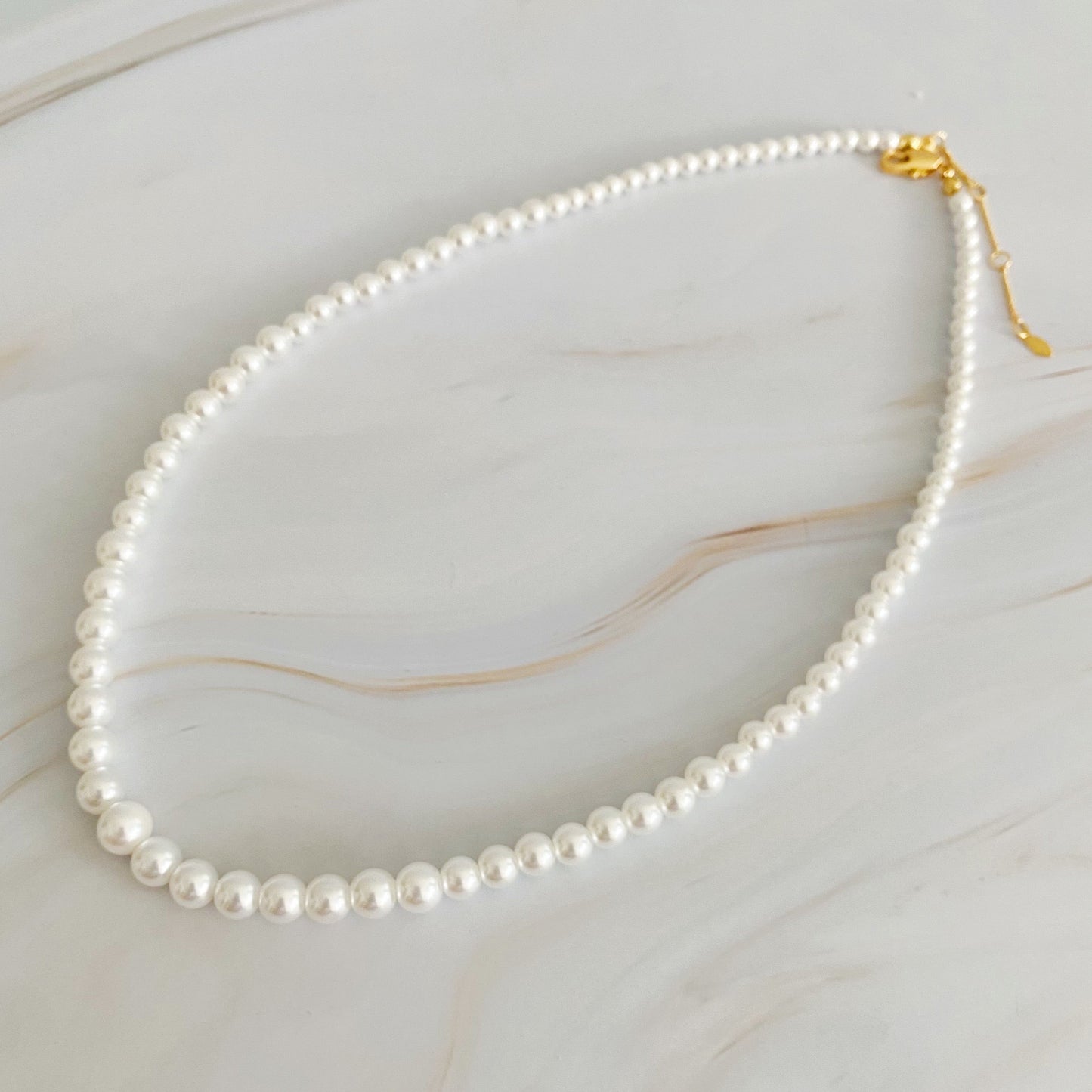 The Queen Liz Graduated Pearl Necklace by Ellison + Young features white pearls with a gold clasp, displayed on a light marble surface.