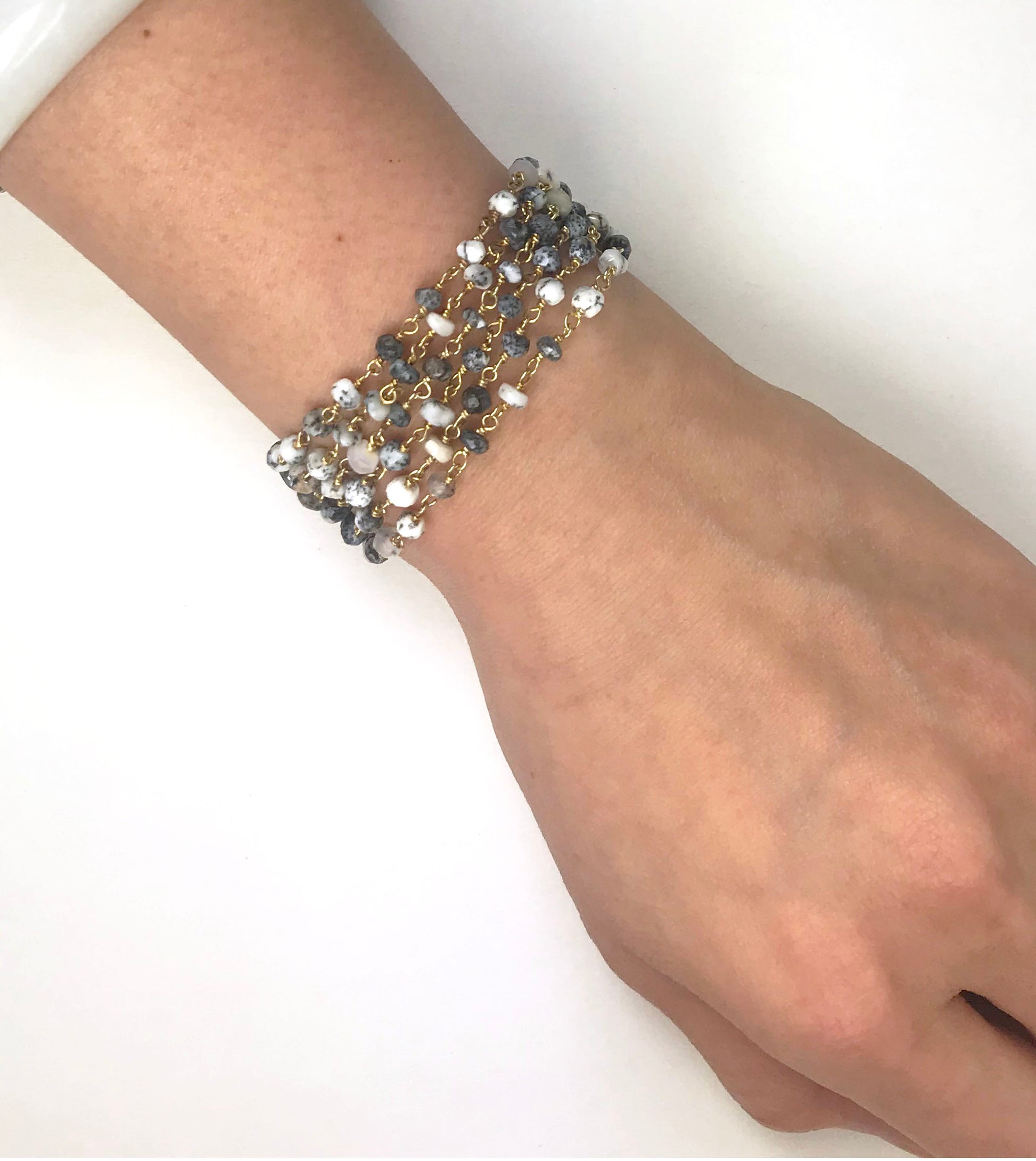 The Dalmatian Opal Bracelet by Gena Myint is a stylish multi-strand accessory with small gray and white beads linked by gold chains, complemented with a gold clasp and heart charm.