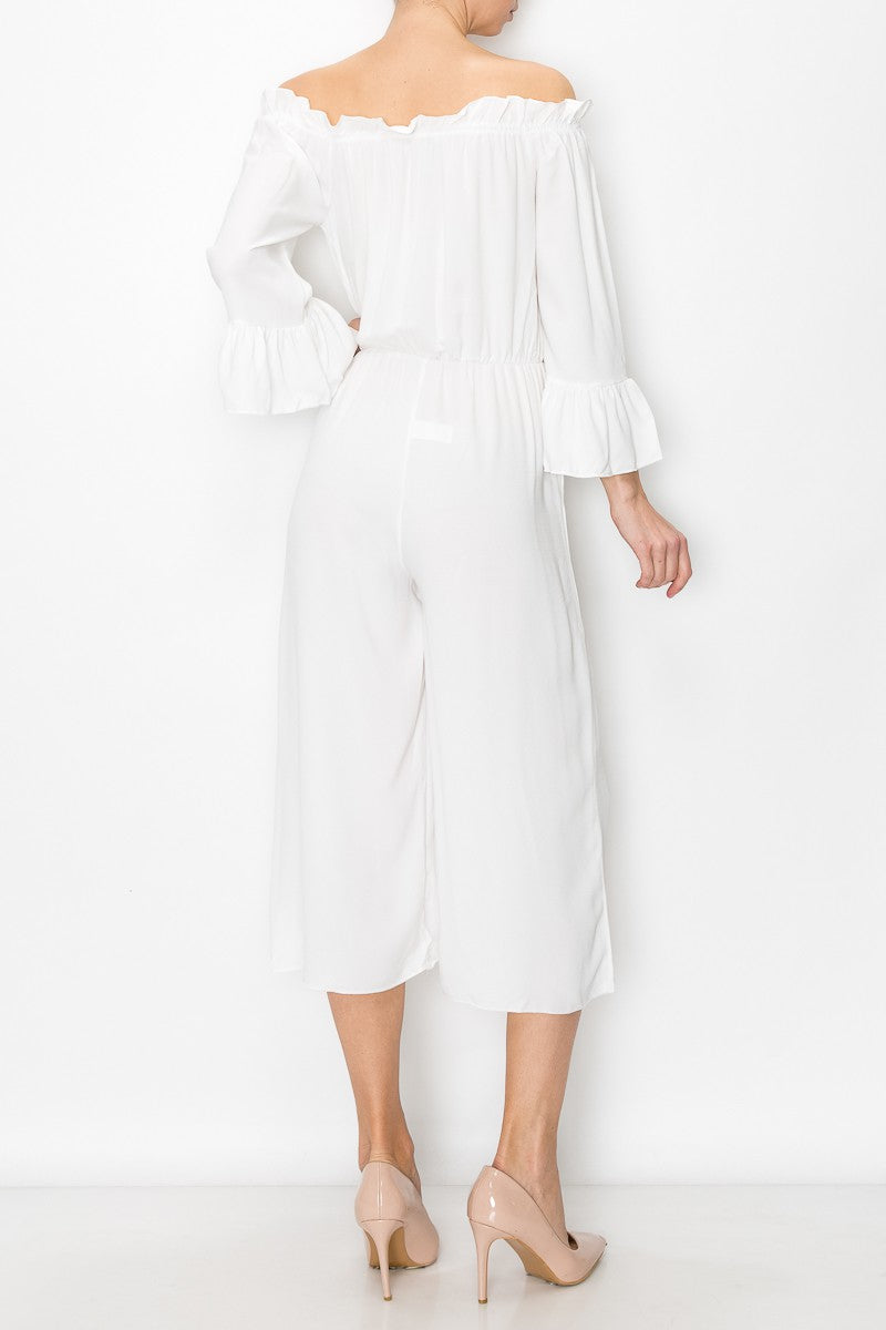 A person models Stylespects Off Shoulder Jumpsuit in white, featuring wide legs, paired with beige heels against a white backdrop.